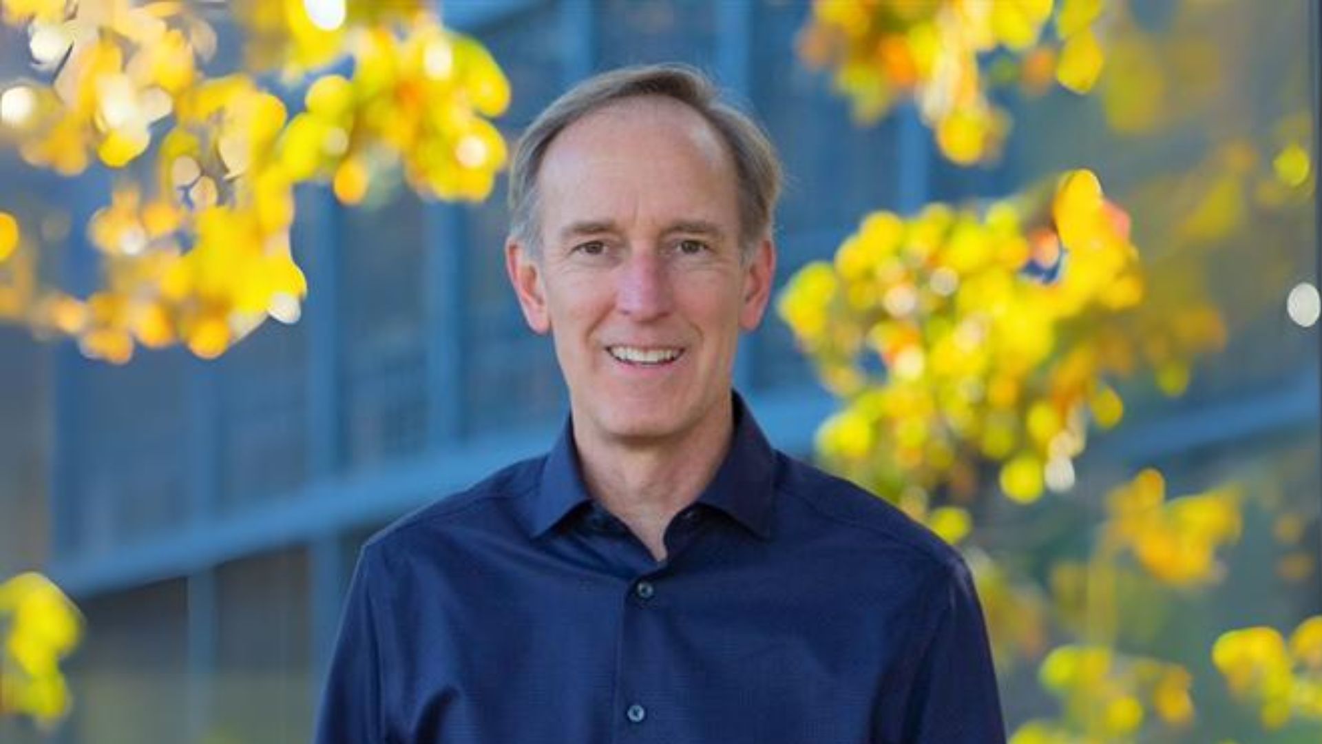 Bishop Tom Berlin standing in front of yellow leaves and a grey building